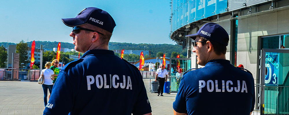 Dwóch funkcjonariuszy policji podczas patrolu w mieście w letni, słoneczny, gorący dzień. Policjanci posiadają umundurowanie dostosowane do warunków pogodowych. Granatowe koszule polo z krótkim rękawem, czapki z daszkiem i okulary przeciwsłoneczne.