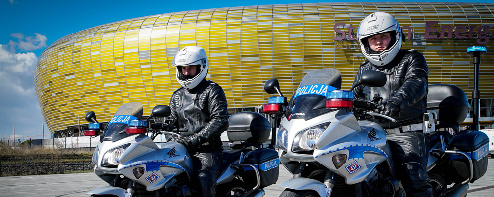 Policjanci na motorach w białych kaskach motorowych i w specjalistycznym umundurowaniu ochronnym do jazdy na motorze w kolorze czarnym. W tle stadion piłkarski w Gdańsku.