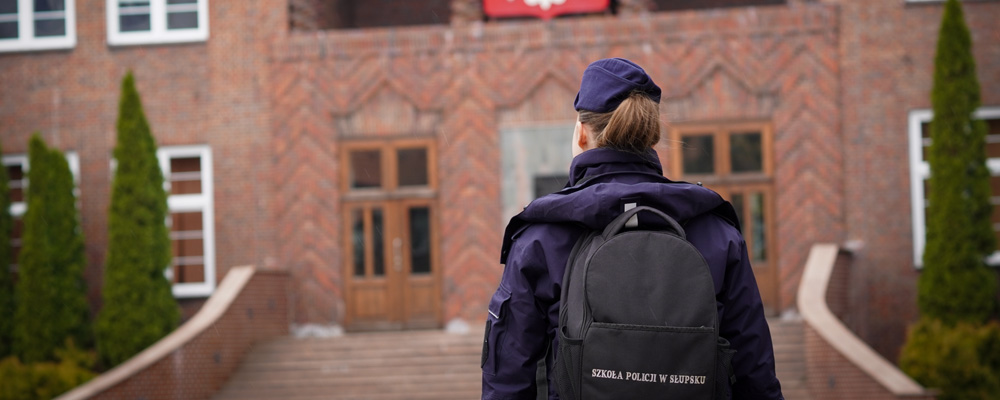 Policjantka z plecakiem mającym napis SZKOŁA POLICJI W SŁUPSKU przed wejściem do budynku szkoły