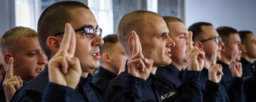 Kandydaci do policji składają ślubowanie. Stoją na baczność w mundurach z uniesioną reką z wyprostowanymi palcami serdecznym i wskazującym i wymawiający rotę przysięgi.