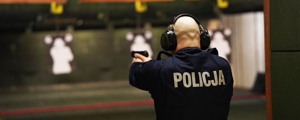 Policjant podczas kursu podstawowego na strzelnicy policyjnej w szkole policji. Strzela do tarczy z pistoletu w pozycji wyprostowanej z nałożonymi słuchawkami ochronnymi na uszy.