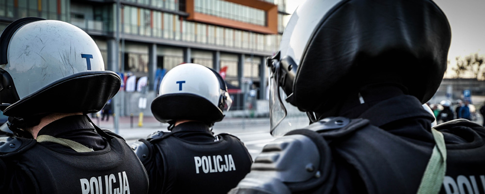 Policjanci Oddziałów Prewencji Policji w umundurowaniu ochronnym wyrózniającym sie białymi chełmami ochronnymiz przyłbicą oraz pancerzem ochronnym na nogi ręce i tułów podczas zabezpieczenia meczu piłkarskiego.