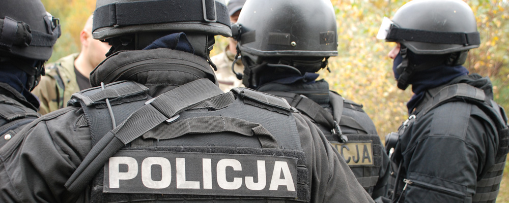 Policjanci Samodzielnego Oddziału Kontrterrorystycznego Policji w chełmach ochronnych i czarnym umundurowaniu przygotowujący się do ćwiczeń. W tle instruktorzy omawiącjący plan ćwiczeń.