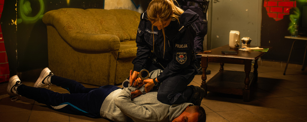 Policjantka aresztuje podejrzanego. Policjantka przytrzymuje leżącego podejżanego kolanem i zakłada mu kajdanki. W tle jej partner zabezpiecza pomieszczenie, w którym dochodzi do aresztowania.