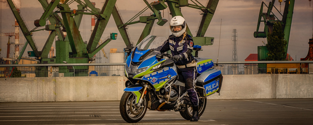 Nowoczesny motor policyjny z nowym malowaniem z siedzącom na nim policjanką. Nowe malowanie radiowowzów i motorów składa się oklein w jaskraych odblaskowych kolorów, żółtegi i niebieskiego. Pojazdy są w kolorze szarym.